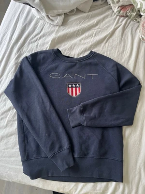 Mörkblå sweatshirt från GANT - Mörkblå sweatshirt från GANT med broderad logga och sköld på bröstet. Klassisk rund halsringning och ribbade muddar vid ärmslut och nederkant. Jätte skön och bra skick