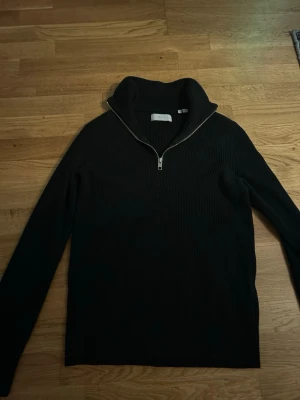 Svart ribbstickad halvzip-tröja - Svart ribbstickad tröja med halvzip och hög krage. Tröjan har långa ärmar och en stilren look som passar perfekt till jeans eller kjol. Materialet känns mjukt och bekvämt, och dragkedjan framtill ger en cool detalj.