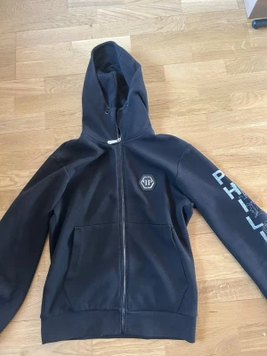 Svart Philipp Plein hoodie med dödskalle - Svart hoodie från Philipp Plein med dragkedja, stor huva och fickor framtill. På bröstet finns en logga och på ryggen en stor dödskalle med texten PHILIPP. Ärmen har också tryckt text. Perfekt för dig som gillar unika och stiliga tröjor. ‼️Som jag visa har den ett väldigt litet hål i innerfickan som man inte kan se utan på tröjan, likadant hål på andra fickan. Sen en väldigt liten fläck på bakre armen som man inte ser på långt håll‼️