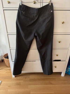Svarta golfbyxor från Puma - Snygga svarta golfbyxor från Puma med raka ben och diskret logga på höften. Byxorna har en klassisk passform, bälteshällor och sidofickor. Perfekta för sportiga aktiviteter eller en aktiv vardag.