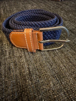 Grisch flätat bälte  - Snyggt mörkblått flätat bälte med detaljer i brunt skinn och silverspänne. Perfekt accessoar för att lyfta din outfit med en klassisk och stilren look. Passar till både jeans och chinos. Väldigt bra kvalité 