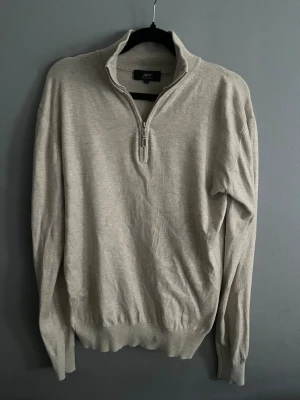 Beige halvzip tröja från Jeff - Snygg beige tröja från Jeff med halvzip och hög krage. Tillverkad i mjukt material och har ribbade muddar vid ärmslut och nederkant. Perfekt för lager på lager och enkel att matcha med olika outfits.