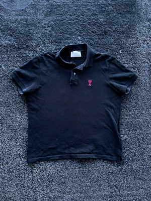 Ami Paris pike - Svart pikétröja från Ami Paris med klassisk krage och knappslå. Tröjan har korta ärmar och en liten röd broderad logga på bröstet. Tillverkad i mjuk bomull som känns skön mot huden. Perfekt för en clean och stilren look. Köpt på NK, storlek M/S, skick 9/10. Pris går att diskutera✅