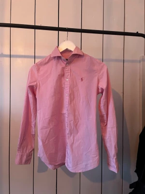Rosa skjorta från Ralph Lauren - Klassisk rosa skjorta från Ralph Lauren med lång ärm och knappar framtill. Skjortan har en broderad logga på bröstet och är tillverkad i bomull. Perfekt för en stilren och avslappnad look. På skjortan står det storlek 2 men de är som 160. Är dock osäker ifall skjortan är redig.