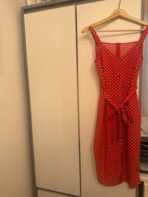 Röd prickig klänning med knytband - Superfin röd klänning med vita prickar, knappar framtill och knytband i midjan. Klänningen har ärmlös design med breda axelband och v-ringning. Perfekt för varma dagar och ger en retrovibe.