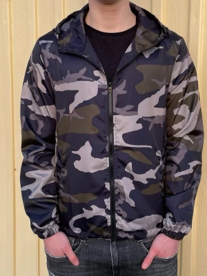 Camo windbreaker - Snygg vindjacka med camouflagemönster i svart, grått och grönt. Jackan har dragkedja framtill, elastiska muddar och en praktisk huva. Perfekt för blåsiga dagar och enkel att matcha med jeans eller streetwear.