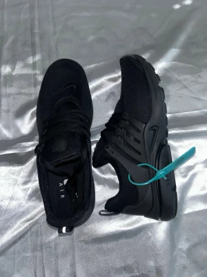 Svarta Nike Air sneakers - Snygga helsvarta Nike Air sneakers med låg profil och klassisk swoosh på sidan. Skorna har en sportig design med mesh- och syntetmaterial, samt en turkos tagg på ena skon för extra detalj. Perfekta för dig som gillar stilrena och bekväma sneakers.