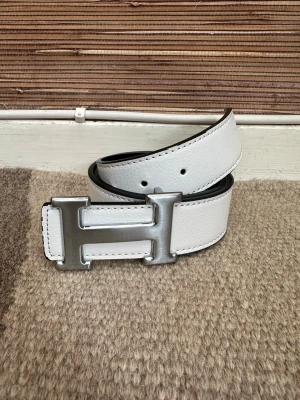 Vitt skärp med H-spänne  - Stilrent vitt skärp i skinn med ett stort, silverfärgat H-spänne. Bältet har svart insida och tydliga sömmar längs kanterna. Perfekt accessoar för att lyfta din outfit.