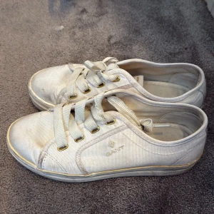 Vita sneakers från Gant st 37 - Klassiska vita sneakers från Gant med lågt skaft och rund tå. Skorna har vita snören, diskret logga på sidan och detaljer i guld vid snörhålen. Tillverkade i textil med gummisula. Perfekta för en clean och stilren look. Lite tvätt så blir de som nya