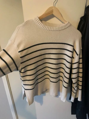 Vit stickad tröja med svarta ränder - Snygg vit stickad tröja med svarta horisontella ränder och ribbad kant nertill. Tröjan har en avslappnad passform, rund halsringning och slitsar i sidorna. Perfekt för en trendig och bekväm look.