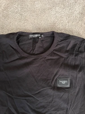 Svart Dolce & Gabbana t-shirt - Snygg svart t-shirt från Dolce & Gabbana med rund hals och en diskret logopatch på bröstet. Tillverkad i mjuk bomull och har en klassisk passform med korta ärmar. Perfekt för dig som gillar stilrena och exklusiva plagg.