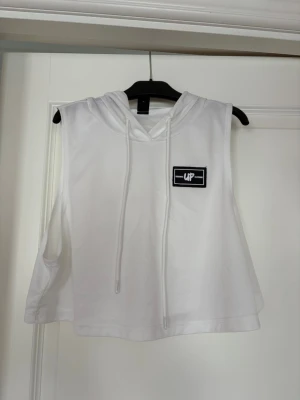 Vit croppad ärmlös hoodie  - Vit croppad ärmlös hoodie med huva och snören. Svart litet märke på bröstet, ’Original UP clothing’. Den är tillverkad i ett lätt, sportigt material som passar perfekt till träning och som är genomskinligt. 