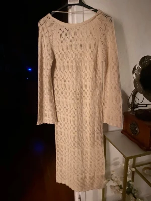 Beige stickad långklänning  - Beige stickad långklänning med lång ärm och vackert hålmönster.  ( kan ändra pris). Klänningen har rund halsringning och är tillverkad i ett mjukt stickat material som ger en mysig känsla. Perfekt för dig som gillar en enkel men stilren look till sommaren och stranden. 