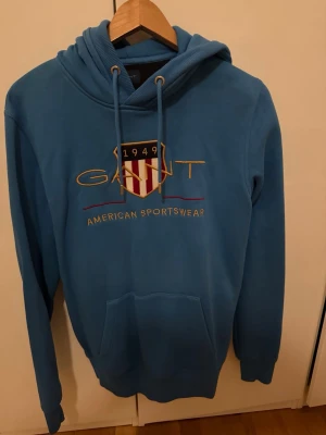 Blå hoodie från Gant med brodyr - Snygg blå hoodie från Gant med broderad logga och texten 'American Sportswear' på bröstet. Klassisk huva med dragsko och stor magficka framtill. Skön och stilren modell som passar perfekt till jeans eller joggers.