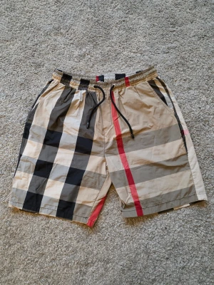 Rutiga shorts med snörning - Snygga shorts med klassiskt rutigt mönster i beige, svart, grått och rött. De har elastisk midja med svart snörning och sidofickor. Perfekta för sommarbaden och ger en stilren vibe.