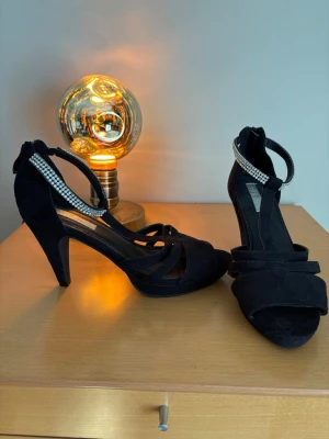 Svarta pumps med strassdetalj - Snygga svarta pumps från Vox Shoes med öppen tå och smala remmar. Klackarna har en elegant strassprydd rem runt vristen som ger extra bling. Skorna är i mockaliknande material och har en klassisk siluett med hög klack.