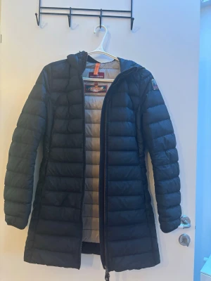 Svart lång dunjacka Parajumpers XS - Säljer en svart lång dunjacka från Parajumpers i superlätt material. Jackan har huva, dragkedja framtill och diskret logga på ärmen. Insidan är ljusgrå och jackan är perfekt för kalla vinterdagar. Snygg och stilren design som håller värmen. Köpt för 5599
