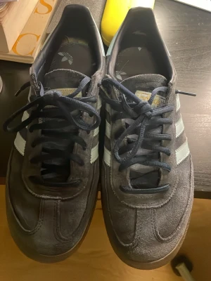 Adidas Spezial marinblå skor - Säljer ett par Adidas Spezial sneakers i marinblå mocka med klassiska ljusblåa ränder på sidorna och gummisula. Skorna har mörkblå snören och gulddetaljen 'SPEZIAL' på sidan. Perfekta för dig som gillar retrostil och streetwear. Dessa har använts väldigt sparsamt och ser nästan nya ut, men de säljs i befintligt skick. De passar till nästan alla kläder och är jättesköna dessutom. Anledningen till att jag säljer dem är endast för att mina fötter har växt lite. Hör av dig vid intresse! 