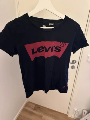 Svart Levi's t-shirt med röd logga - Klassisk svart t-shirt från Levi's med den ikoniska röda loggan tryckt på bröstet. T-shirten har rund halsringning och korta ärmar. Tillverkad i mjuk bomull för en skön känsla. Perfekt till jeans eller shorts för en avslappnad look.