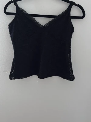 Svart spetslinnetopp med v-ringning - Snygg svart linnetopp med tunna axelband och v-ringad hals. Toppen är helt täckt av spets och har en feminin look. Perfekt att styla med jeans eller kjol för en trendig outfit.