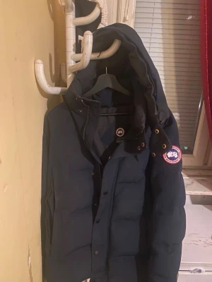 Mörkblå dunjacka från Canada Goose - Säljer en mörkblå dunjacka från Canada Goose med avtagbar huva och pälskant. Jackan har klassisk patch på ärmen, dragkedja och knappar framtill samt flera fickor. Perfekt för kalla vinterdagar och riktigt varm.