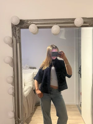 Mörkblå kortärmad kofta med spänne - Snygg mörkblå kofta med korta ärmar och ett dekorativt spänne vid halsen. Koftan har en öppen front och är tillverkad i ett mjukt, stickat material. Perfekt att slänga över en topp eller som på bilden med bara basic linne,livar upp outfiten!