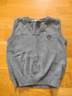 Grå stickad väst med broderad logga - Klassisk grå stickad väst med v-ringning och ribbade kanter från Lindex i storlek 86/92. På bröstet finns en broderad blå logga som ger en preppy känsla. Perfekt att bära över en skjorta för en stilren look.