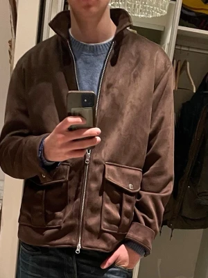mockajacka från Human Scales NY - Helt ny snygg brun suedejacka från Human Scales, modell Seth. Jackan har två stora fickor med lock framtill, dragkedja och en hög krage. Insidan är fodrad med blått tyg som känns lyxigt. Perfekt för dig som vill ha en stilren och trendig jacka.