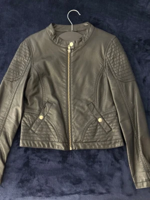 Svart skinnjacka för barn - Svart skinnjacka med guldfärgade dragkedjor. Kort modell och rundad krage. Storlek 158.