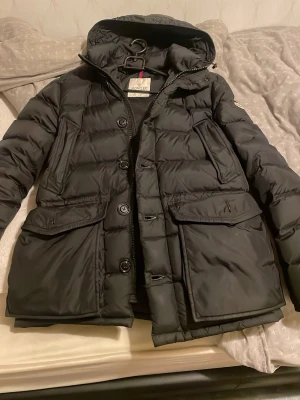 Svart dunjacka från Moncler med huva - Snygg svart dunjacka från Moncler med huva och stora fickor framtill. Jackan har både dragkedja och knappar, quiltad design och klassisk Moncler-logga på ärmen. Perfekt för kalla dagar och riktigt stilren look.