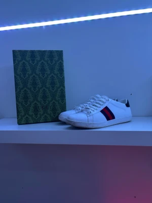 Gucci vita sneakers med blå och röd detalj - Säljer ett par klassiska vita sneakers från Gucci med blå och röd rand på sidan och mörkblå häl med Gucci-logga i silver. Skorna har låga skaft, vita snören och är tillverkade i läder. Perfekta för dig som vill ha en clean och lyxig look.