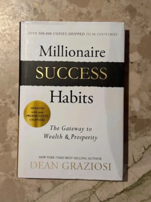 Millionaire Success Habits bok - En inspirerande bok med vit och svart framsida och guldfärgade detaljer, skriven av Dean Graziosi. Titeln 'Millionaire Success Habits' står i stora bokstäver och boken handlar om vanor för framgång och välstånd. Perfekt för dig som vill utvecklas och nå nya mål.