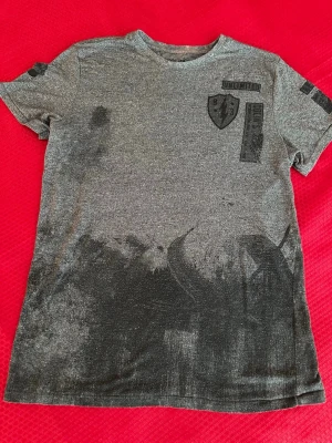 Grå t-shirt med tryck och patch M - Säljer en gråmelerad t-shirt med svart tryck och patchdetaljer på bröstet och ärmarna. T-shirten har rund hals och korta ärmar. Materialet känns mjukt och skönt, troligen bomull eller bomullsblandning. Perfekt för en avslappnad streetwear-look.