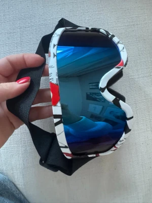 Blå spegelglas skidglasögon - Coola skidglasögon med blått spegelglas och vit ram med svarta och röda detaljer. Justerbart svart band för perfekt passform. Perfekta för dig som vill sticka ut i backen med en sportig look. Har bundlerabatt vid köp av fler varor🥰