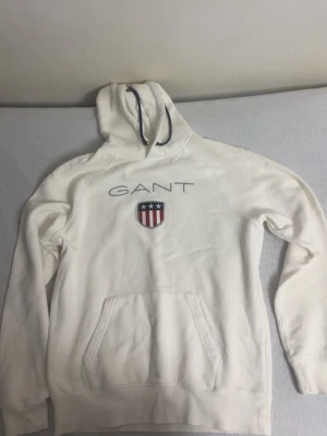 Vit hoodie från GANT med logga - Säljer en vit hoodie från GANT med broderad logga och amerikanskt sköldmärke på bröstet. Hoodien har dragsko i huvan och en stor magficka. Tillverkad i mjukt bomullsmaterial och har ribbade muddar vid ärmar och nederkant. Perfekt för en avslappnad och stilren look.