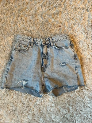 Blå denimshorts med råa kanter - Säljer ett par klassiska blå denimshorts med hög midja och råa, fransiga kanter. Shortsen har en liten slitning framtill och femficksdesign. Perfekta för sommaren och ger en avslappnad vibe.