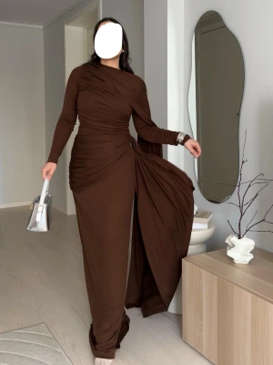 Brun långklänning med drapering - Säljer en elegant brun långklänning med draperade detaljer och lång ärm. Klänningen har en asymmetrisk design med tyg som faller från höften och markerad midja. Perfekt för dig som vill sticka ut med en unik och stilren look.