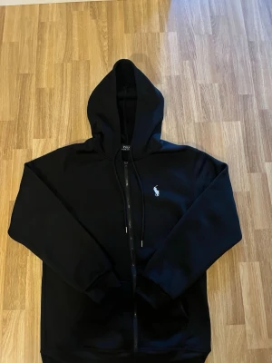 Svart hoodie från Polo Ralph Lauren - Svart hoodie med dragkedja från Polo Ralph Lauren. Klassisk design med huva och justerbara snören samt vit broderad logga på bröstet. Perfekt för en avslappnad och stilren look.