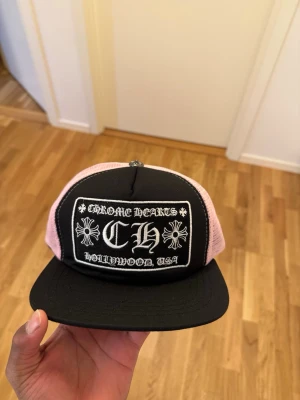 CH keps svart/rosa - Säljer en snygg CH keps med svart front och rosa mesh på sidorna. Kepsen har en rak skärm och ett stort broderat märke framtill med texten  Hollywood, USA' och CH-logga. Justerbar baktill för perfekt passform.
