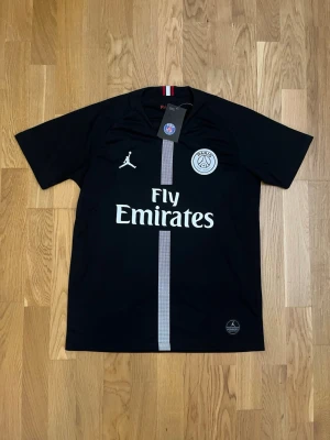 Svart PSG matchtröja Jordan x Nike - Snygg svart PSG matchtröja i samarbete med Jordan och Nike. Tröjan har korta ärmar, vit PSG-logga och Jordan-logga på bröstet samt Fly Emirates-tryck framtill. Vertikal grå rand i mitten och röd-blå detalj i nacken. Tillverkad i lätt och ventilerande material.