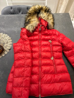 Röd dunjacka med fuskpäls från Tiffosi - Snygg röd dunjacka från Tiffosi med avtagbar huva och fuskpäls. Jackan har quiltad design, dragkedja framtill och två snedställda fickor med dekorativa dragkedjor. Perfekt för kalla vinterdagar och riktigt varm.