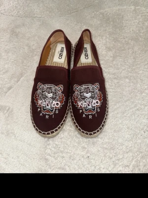 Bordeaux Kenzo ballerinaskor med tiger - Snygga bordeaux ballerinaskor från Kenzo med ikoniskt broderat tigermotiv och texten 'Paris' framtill. Skorna är i canvas med naturfärgad sula i flätad jute och har en klassisk slip-on modell. Perfekta för dig som gillar statement-detaljer och streetstyle.