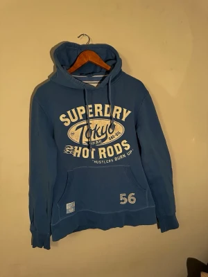Blå Superdry hoodie Tokyo Hot Rods - Blå hoodie från Superdry med tryck i vitt och beige framtill där det står 'Superdry Tokyo Hot Rods'. Klassisk känguruficka, dragsko i huvan och tryckt siffra 56 på magen. Skön och avslappnad passform, perfekt till jeans eller joggers.