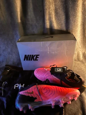 Nike Phantom gx Elite  - Säljer ett par Nike Phantom Gx Elite F fotbollsskor i en snygg rosa och svart färg. Skorna har en strumpliknande passform, avancerad textur för bättre bollkontroll och en lätt yttersula med fasta dobbar för spel på naturgräs. Perfekta för dig som vill sticka ut på planen.