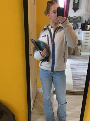 Vit fleecejacka från Patagonia - Mysig vit fleecejacka från Patagonia med blå dragkedja och detaljer i orange. Jackan har hög krage och lång ärm, perfekt vår eller höstjackan, funkar även som mellan lager på tex afterski. Sparsamt änvänd ca 5 gånger 💗💗