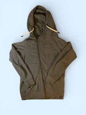 Grå zip hoodie från Replay - Snygg grå hoodie från Replay med dragkedja framtill och stor huva med vita snören. Stora svarta bokstäver 'REPLAY' på ryggen och diskret logga på bröstet. Tillverkad i mjukt bomullsmaterial och har två fickor fram. Perfekt för en avslappnad streetwear-look.