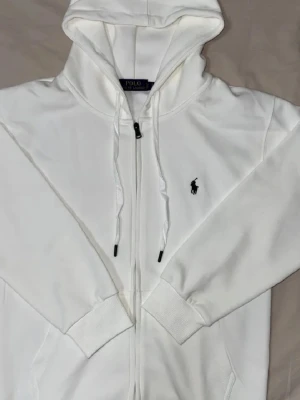 Vit hoodie från Polo Ralph Lauren - Snygg vit hoodie med dragkedja från Polo Ralph Lauren. Klassisk design med svart broderad logga på bröstet, justerbar huva med snören och ribbade muddar. Perfekt för en clean och stilren look.