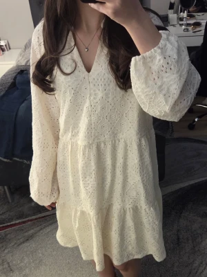 Vit spetsklänning med v-ringning - Romantisk vit klänning med v-ringning och långa ärmar. Klänningen har broderade spetsdetaljer och en luftig passform med volang nertill. Perfekt för sommarens alla tillfällen.