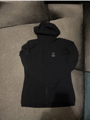 Svart vindjacka från Haglöfs - Säljer en svart vindjacka  från Haglöfs med dragkedja framtill och justerbar huva. Jackan  har diskret logga på bröstet och två sidofickor. Perfekt för dig som gillar enkel och stilren design. Materialet känns mjukt och bekvämt.