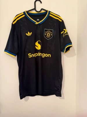 Manchester United svart matchtröja - Snygg svart Manchester United matchtröja från Adidas med gula och blå detaljer. Tröjan har tryck med Snapdragon-logga, klubbmärke och nummer 10 med namnet Cunha på ryggen. Korta ärmar och sportig passform, perfekt för fotboll eller träning.